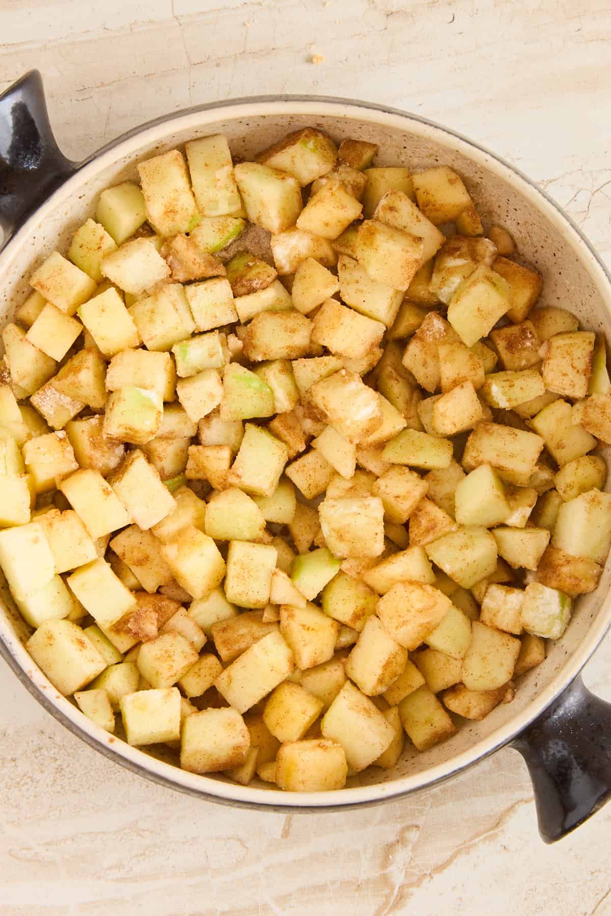 Chopped apples coated with cinnamon and sugar in a large pot, ready to be cooked, seen from above on a light-colored surface.
