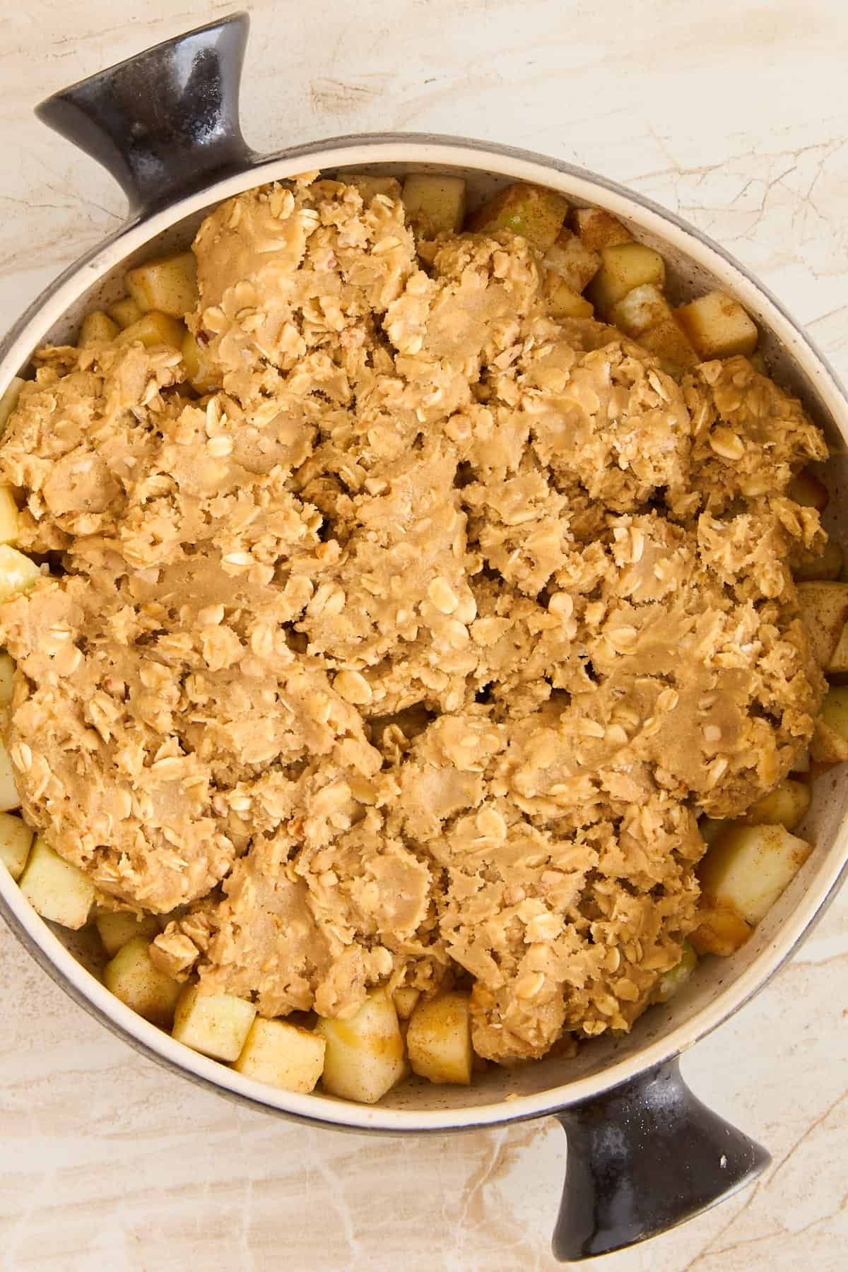 A round baking dish filled with apple chunks, topped with an uneven layer of oat crumble, ready to be baked. The dish has two black handles, and sits on a light-colored countertop.