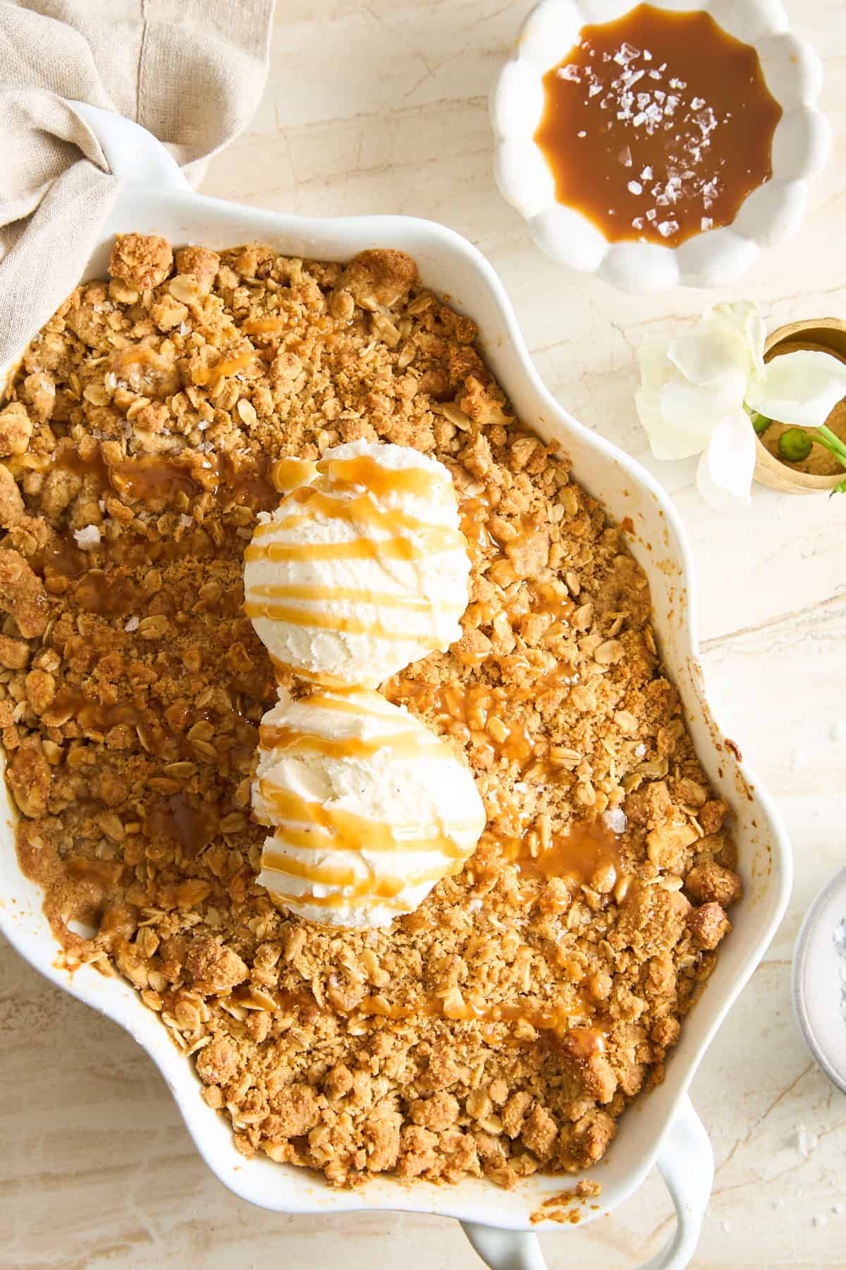 A baked apple crisp topped with two scoops of vanilla ice cream and caramel sauce in a white dish. Next to it is a bowl of caramel sauce sprinkled with sea salt and a white flower on a marble surface.