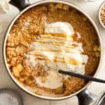 A round baking dish filled with apple crisp topped with melting vanilla ice cream and drizzled with caramel sauce. A spoon rests in the dish. Surrounding the dish are caramel sauce, nuts, a striped cloth, and flowers.