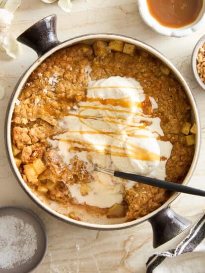 A round baking dish filled with apple crisp topped with melting vanilla ice cream and drizzled with caramel sauce. A spoon rests in the dish. Surrounding the dish are caramel sauce, nuts, a striped cloth, and flowers.