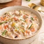 A bowl of creamy soup filled with carrots, celery, chicken, and wild rice, garnished with fresh rosemary and chives, sits on a light table next to gold spoons, with a white flower and another bowl in the background.
