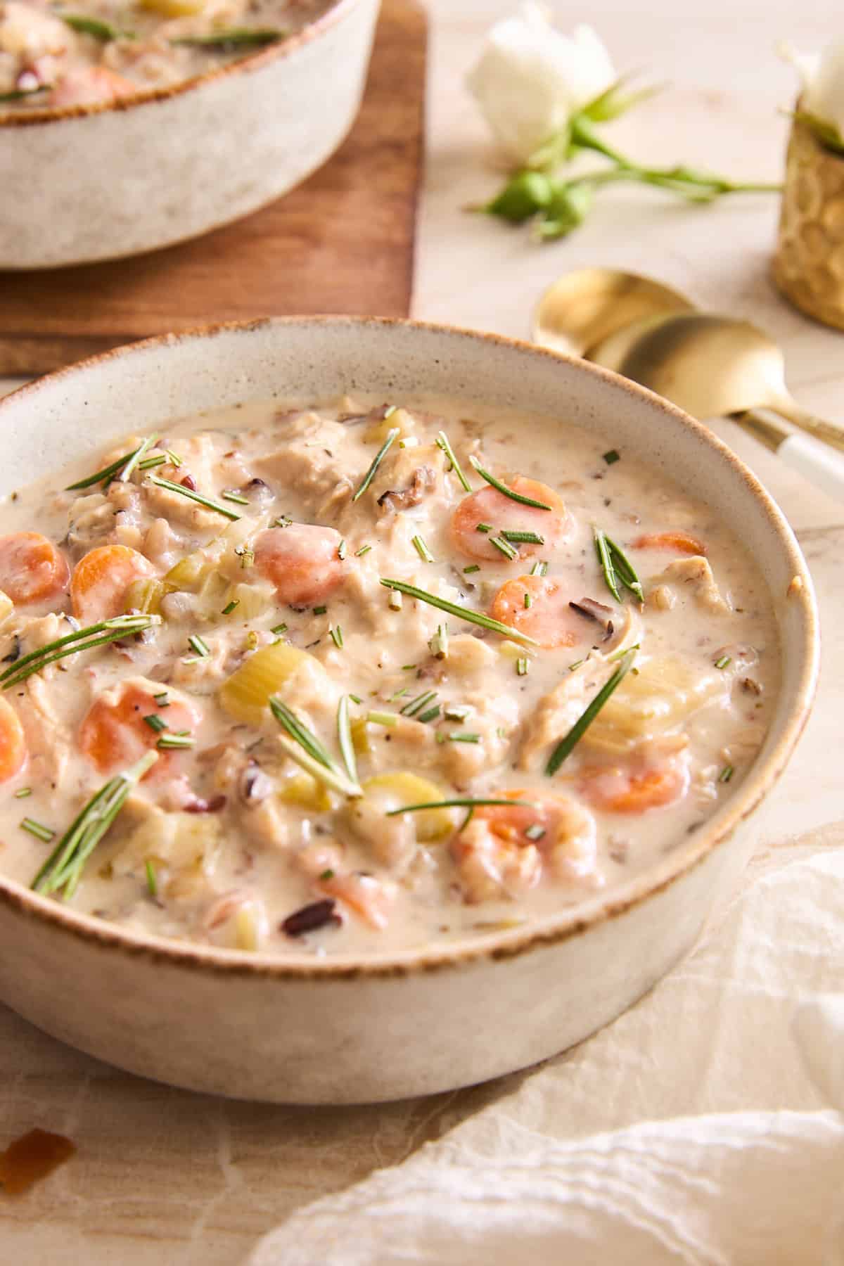 A bowl of creamy soup filled with carrots, celery, chicken, and wild rice, garnished with fresh rosemary and chives, sits on a light table next to gold spoons, with a white flower and another bowl in the background.