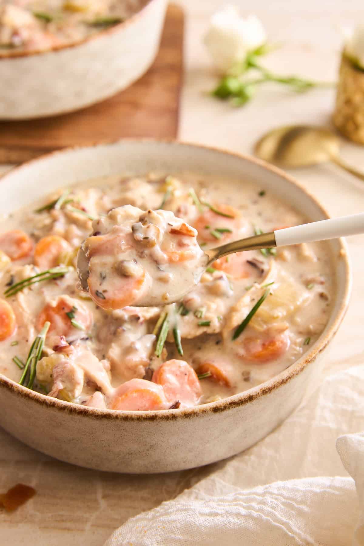 A close-up of a bowl of creamy soup with visible chunks of carrots, mushrooms, and herbs. A spoon is lifting a hearty portion above the bowl. The background includes a cloth napkin and blurred table setting.