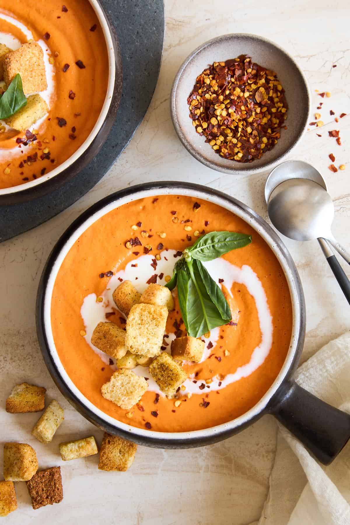 A bowl of creamy tomato soup topped with croutons, fresh basil, and a swirl of cream, beside a bowl of red pepper flakes and scattered croutons, with a spoon and napkin on a light surface.