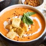 A bowl of creamy tomato soup topped with croutons, a swirl of cream, fresh basil leaves, and a sprinkle of red pepper flakes, with a small dish of red pepper flakes in the background.