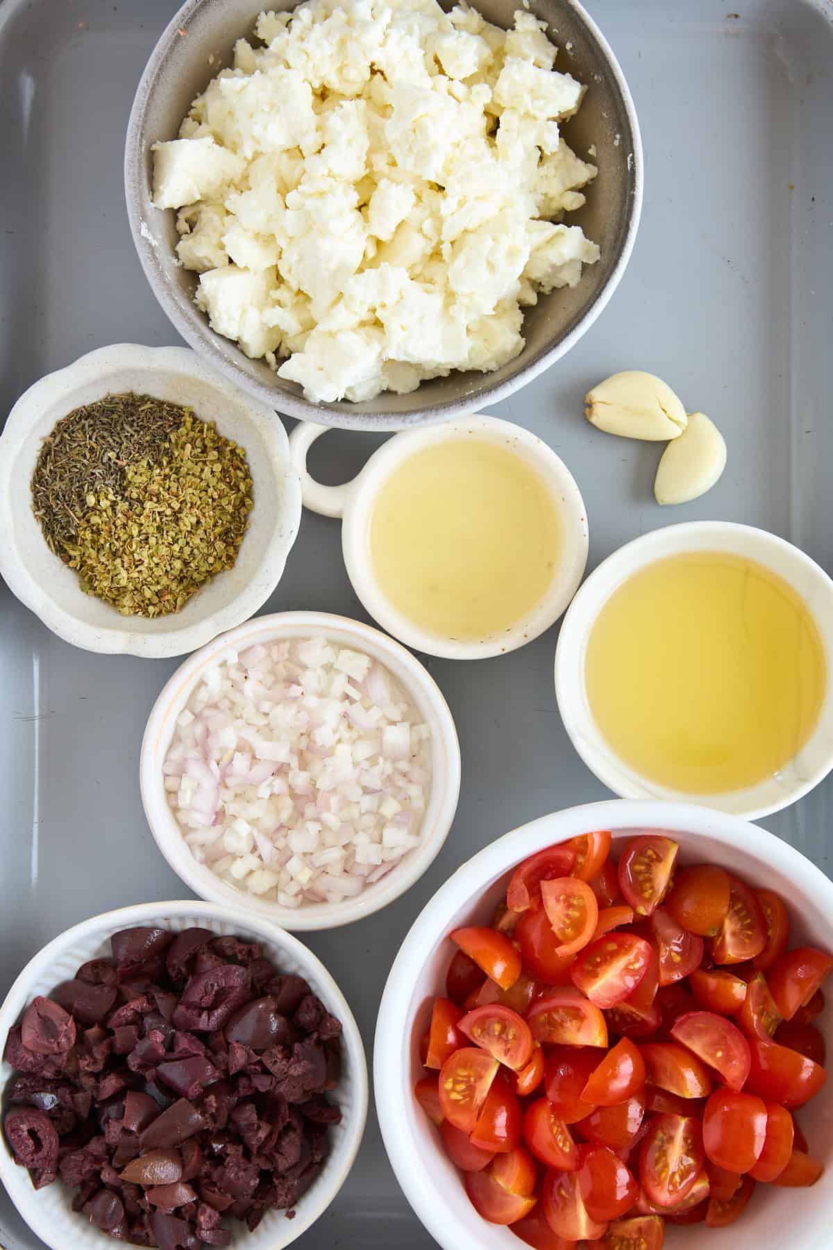 A tray with bowls containing crumbled cheese, halved cherry tomatoes, chopped shallots, sliced black olives, dried herbs, olive oil, white wine, and two cloves of garlic.