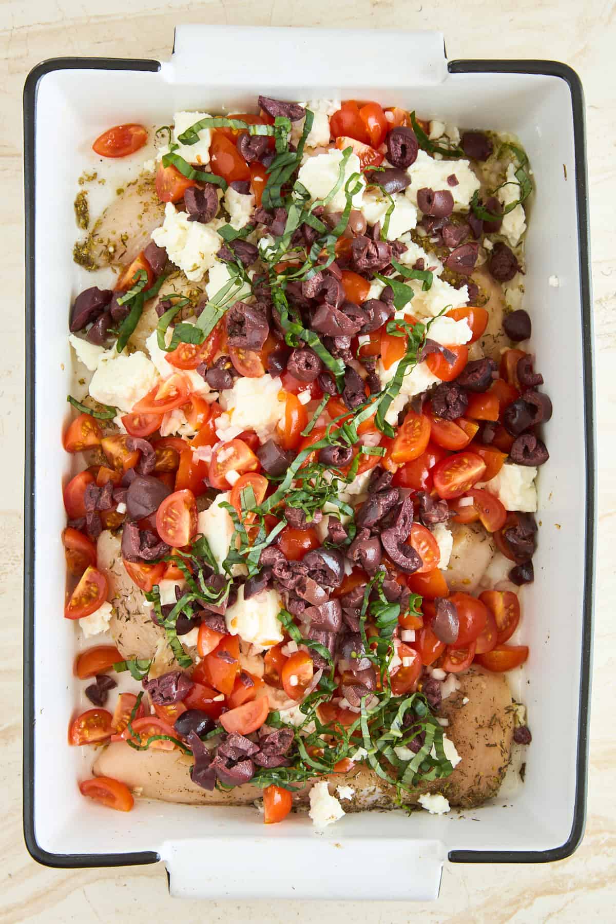 A baking dish filled with raw chicken breasts topped with halved cherry tomatoes, crumbled feta cheese, sliced black olives, and fresh basil strips, ready to be cooked.