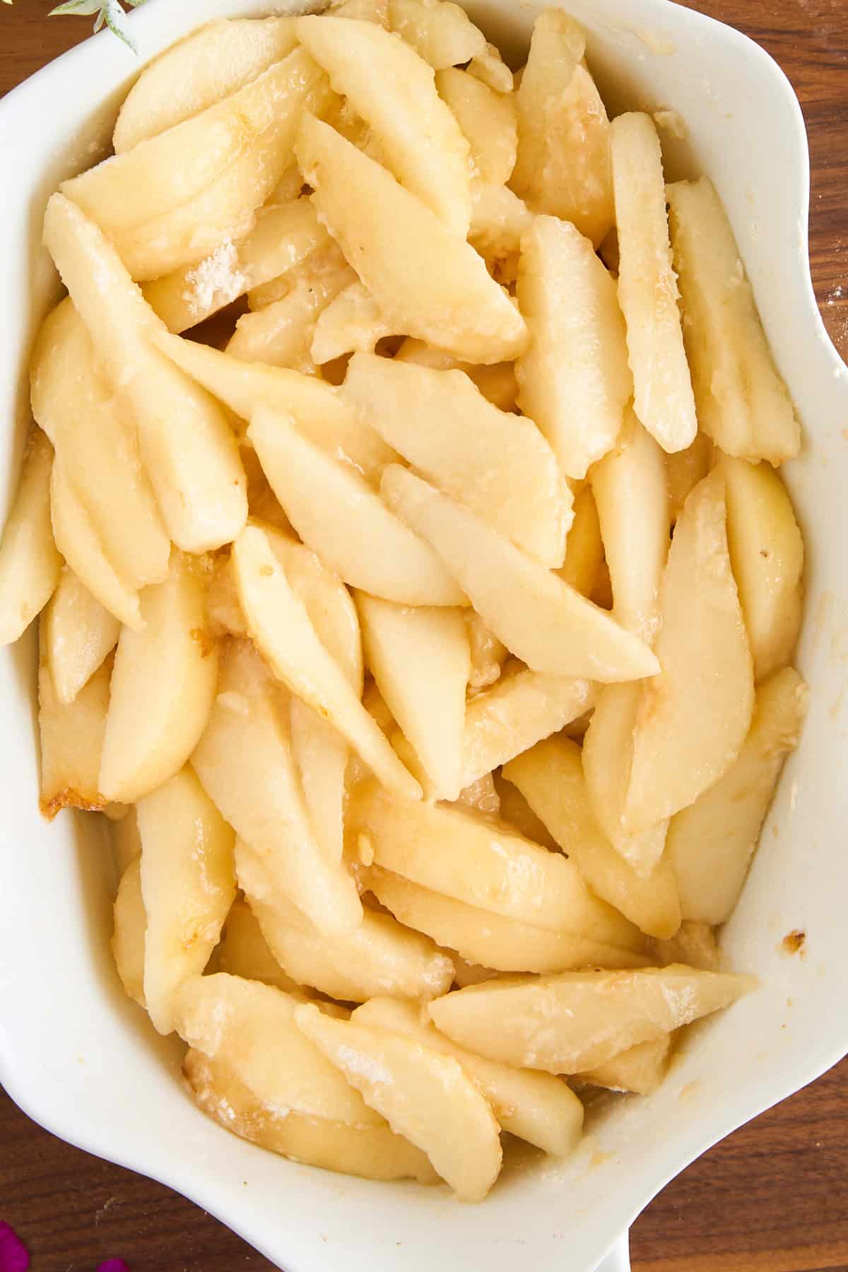 Sliced fresh pears coated with sugar and arranged in a white ceramic baking dish, ready for baking. The pears have a glossy appearance and are evenly distributed in the dish.