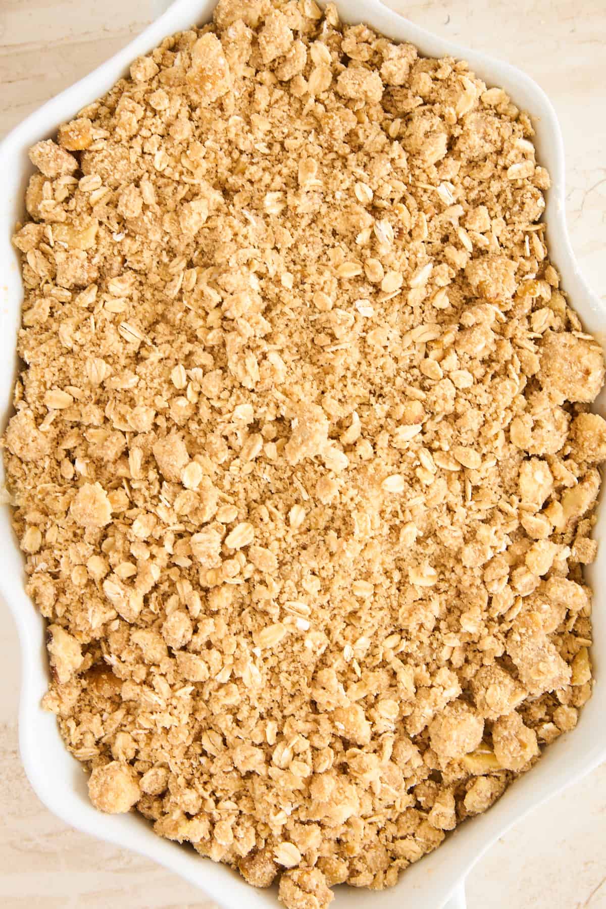 A close-up of an unbaked oat crumble topping in a white baking dish, showing a coarse mixture of oats, flour, butter, and sugar.