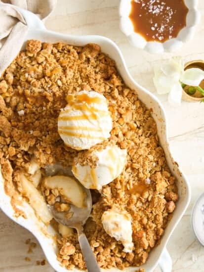 A baking dish filled with apple crisp topped with three scoops of vanilla ice cream and caramel drizzle. A spoon rests inside, and a bowl of caramel sauce sits nearby on a light-colored table.