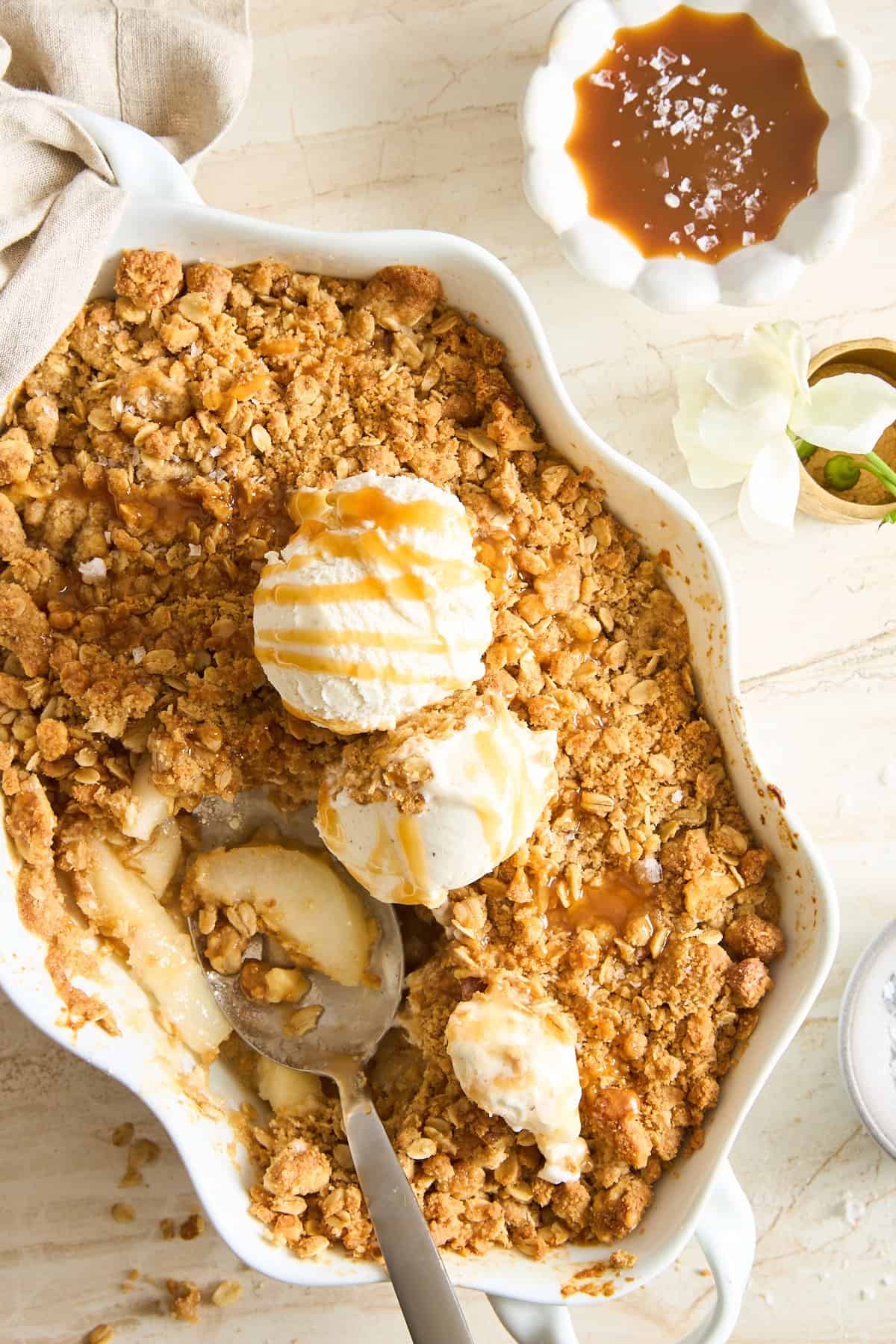 A baking dish filled with apple crisp topped with three scoops of vanilla ice cream and caramel drizzle. A spoon rests inside, and a bowl of caramel sauce sits nearby on a light-colored table.
