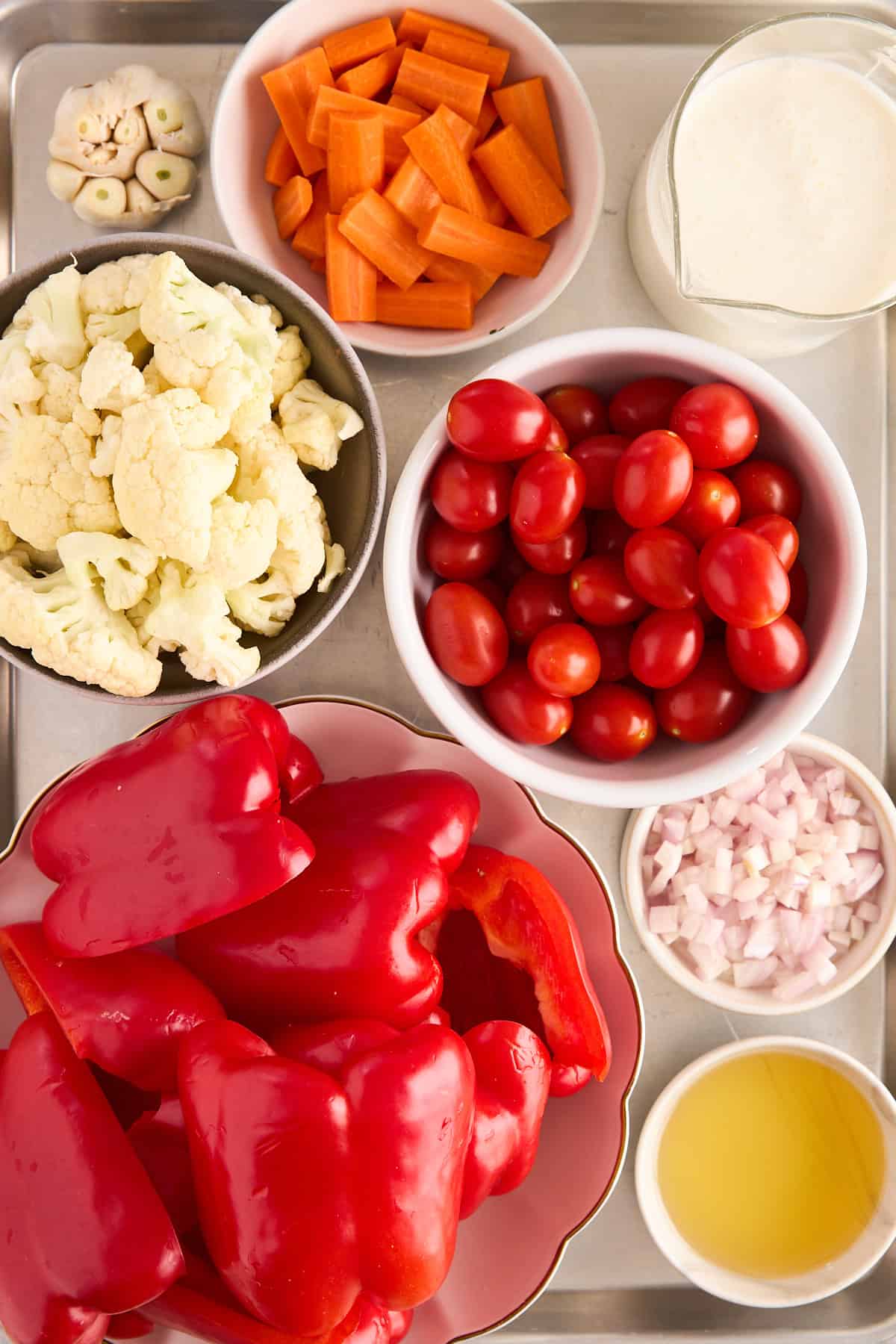 A tray with bowls of fresh vegetables and ingredients: chopped carrots, cauliflower florets, grape tomatoes, red bell peppers, diced shallots, a halved garlic bulb, a glass of cream, and a dish of olive oil.