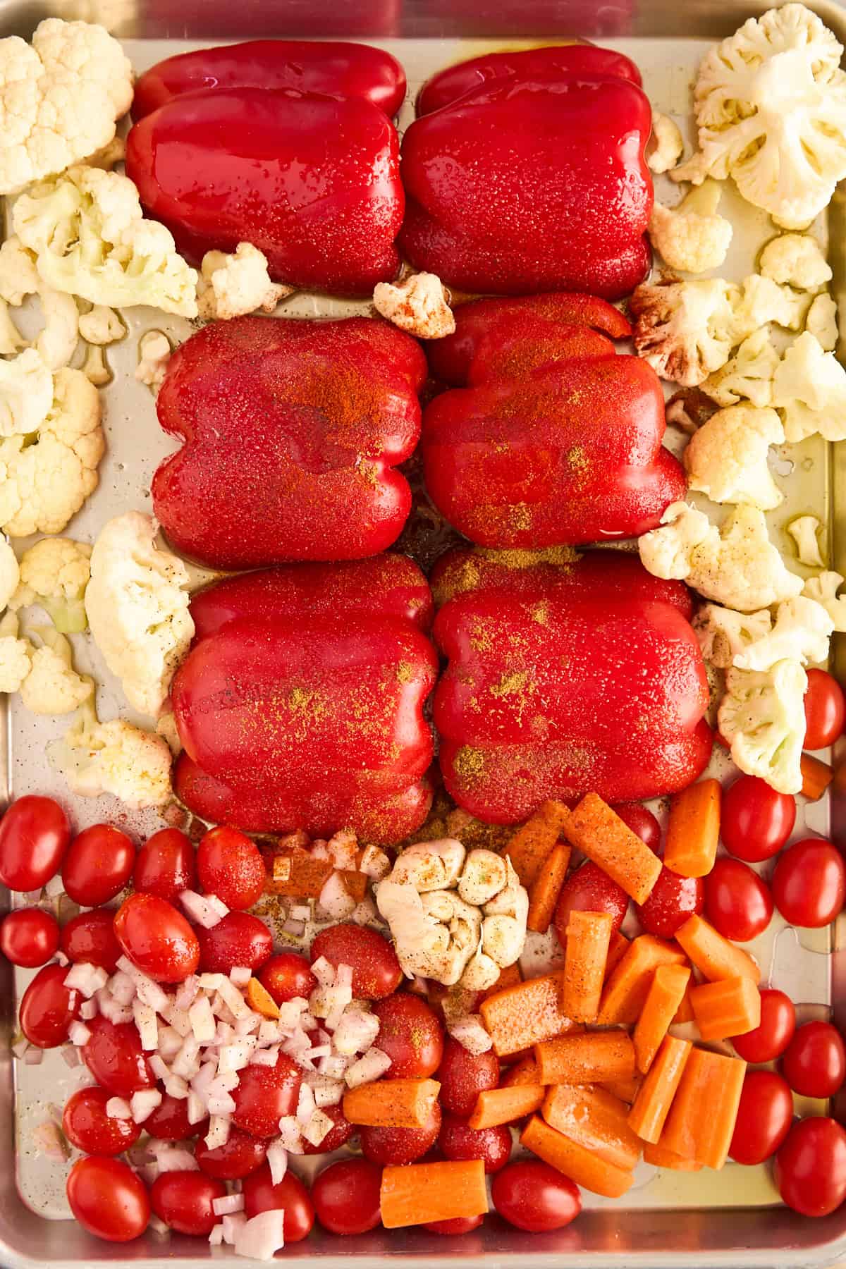 A baking tray filled with red bell peppers, cauliflower florets, grape tomatoes, chopped onions, carrot pieces, and garlic, all seasoned and arranged in neat sections, ready for roasting.