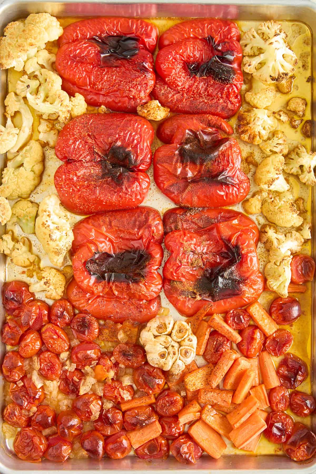 A baking sheet with roasted red bell peppers, cauliflower florets, cherry tomatoes, whole garlic bulbs, and chopped carrots, all glistening with oil and slightly charred from roasting.