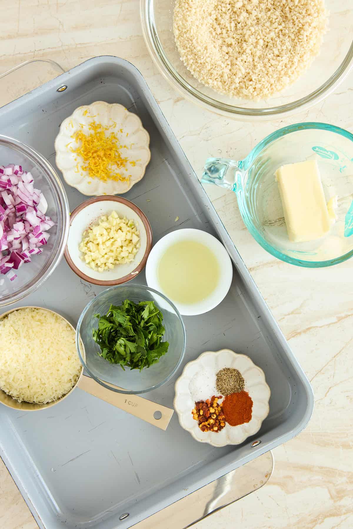 A tray with bowls of chopped red onion, minced garlic, lemon zest, grated cheese, chopped parsley, oil, and spices. Nearby are a bowl of breadcrumbs and a measuring cup with butter on a light countertop.