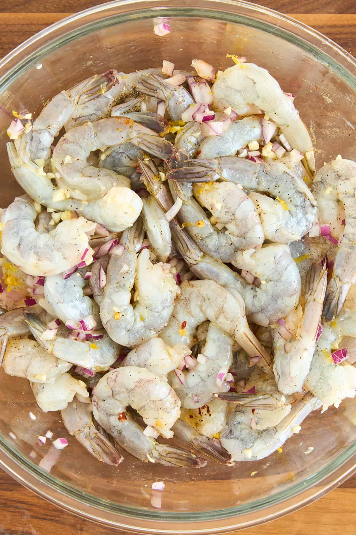 Raw shrimp marinating in a glass bowl with chopped red onions, minced garlic, lemon zest, and spices on a wooden surface.