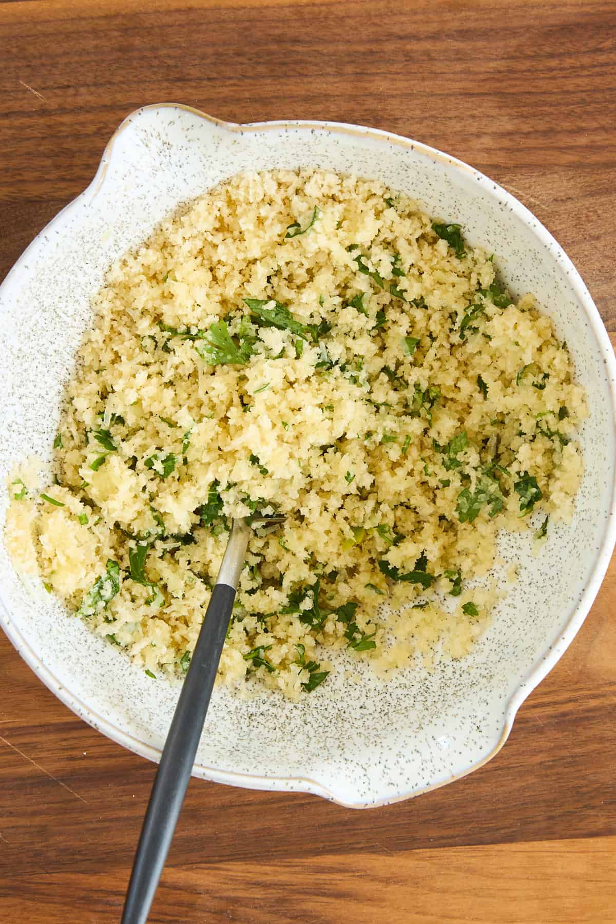 A white speckled bowl filled with a mixture of breadcrumbs, chopped herbs, and grated cheese sits on a wooden surface. A spoon rests inside the bowl.