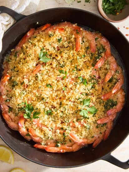 A skillet filled with baked shrimp arranged in a circle, topped with golden breadcrumb mixture and sprinkled with fresh parsley. Lemon wedges and a bowl of chopped herbs are beside the skillet on a light surface.