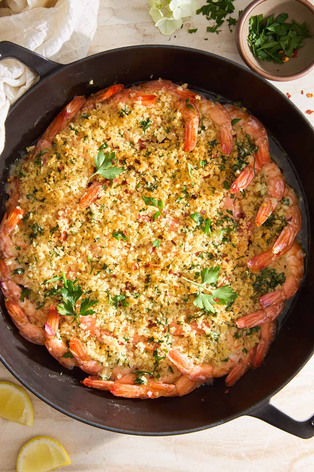 A skillet filled with baked shrimp arranged in a circle, topped with golden breadcrumb mixture and sprinkled with fresh parsley. Lemon wedges and a bowl of chopped herbs are beside the skillet on a light surface.