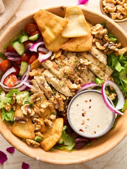 A wooden bowl filled with salad greens, grilled chicken slices, cherry tomatoes, cucumber, red onion, kalamata olives, walnuts, pita chips, and a small dish of dressing.