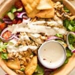 A wooden bowl filled with salad greens, cherry tomatoes, cucumber, red onion, pita chips, walnuts, olives, and drizzled with creamy dressing. A small bowl of extra dressing sits inside the salad.