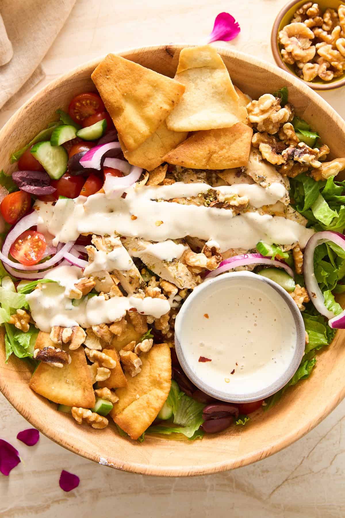 A wooden bowl filled with salad greens, cherry tomatoes, cucumber, red onion, pita chips, walnuts, olives, and drizzled with creamy dressing. A small bowl of extra dressing sits inside the salad.