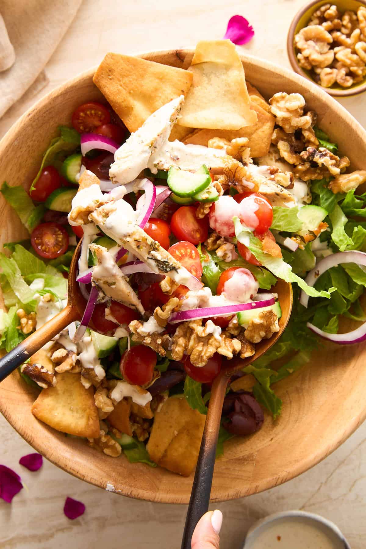 A wooden bowl of fresh salad with lettuce, cherry tomatoes, cucumber, red onion, walnuts, pita chips, and creamy dressing, being served with wooden salad tongs.