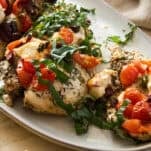 Herb-seasoned baked chicken breasts topped with chopped tomatoes, olives, feta cheese, and fresh basil, served on a white oval plate, with a lemon wedge visible in the corner.