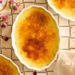 An oval dish of crème brûlée with a caramelized sugar crust sits on a tiled surface, surrounded by scattered dried rosebuds and two partial dishes of crème brûlée. A beige cloth is partially visible in the corner.