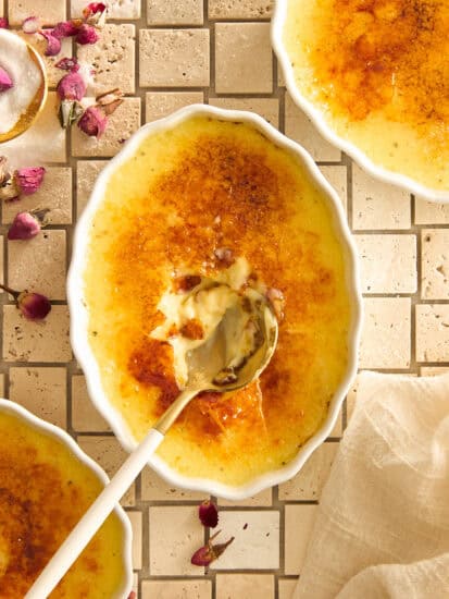 Overhead view of a crème brûlée dish with a caramelized sugar top cracked open by a spoon. Other dishes and scattered dried rosebuds are visible on a cream-colored tile surface. A beige napkin is to the side.