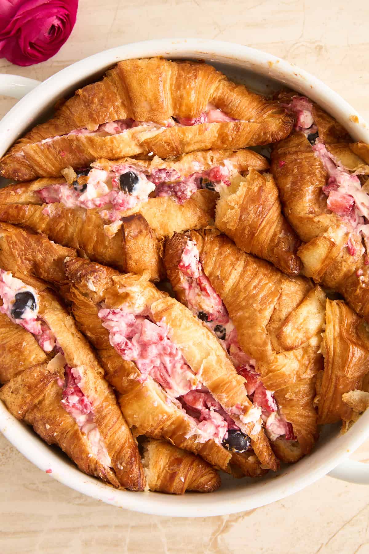 A dish filled with golden croissants, sliced and stuffed with a creamy mixture containing berries. A pink rose is placed at the top left corner of the image on a light background.