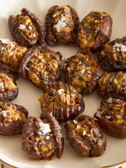 A plate of stuffed dates topped with drizzled chocolate and flakes of sea salt, arranged in a circular pattern on a cream-colored dish.