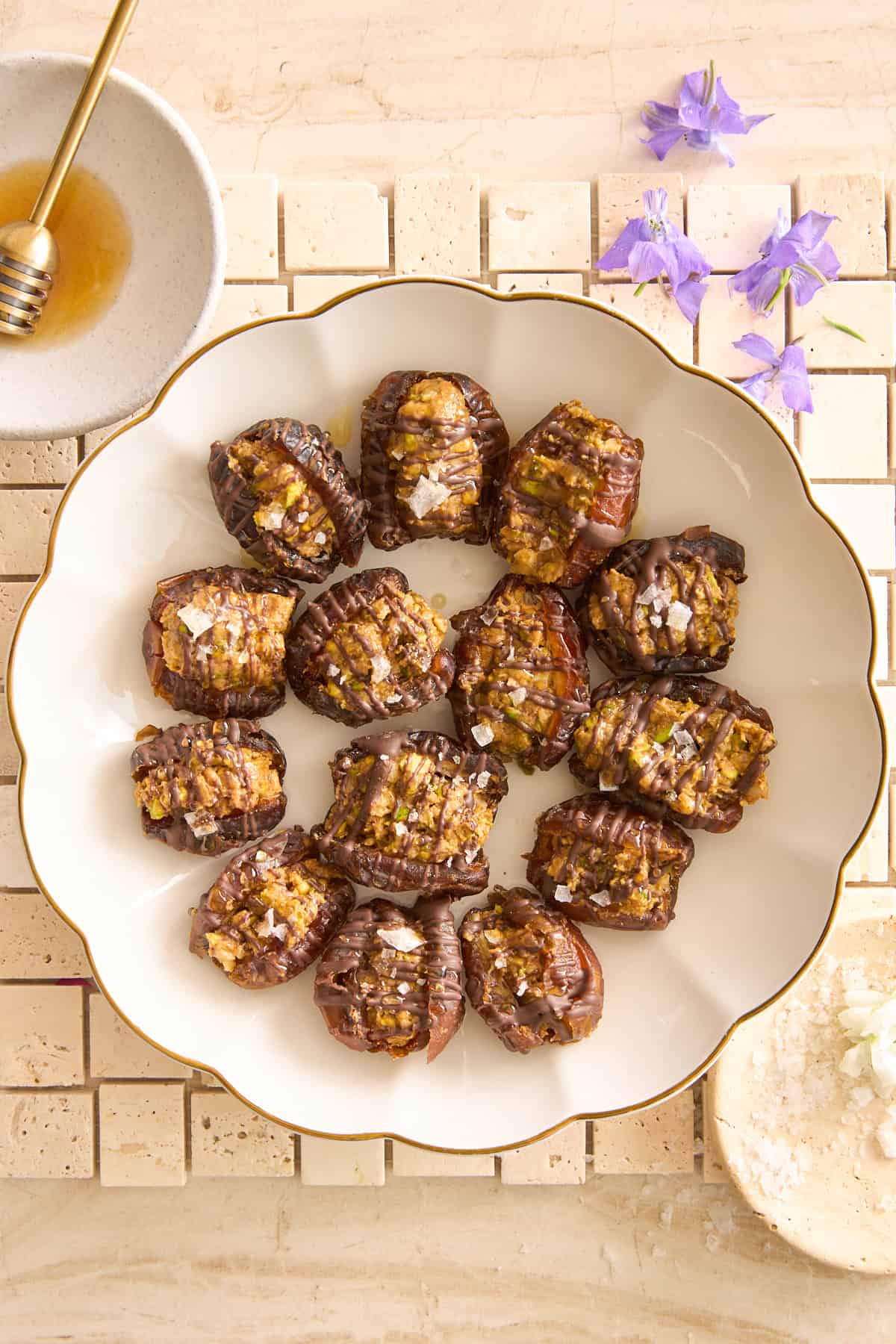 A scalloped white plate filled with sliced, roasted eggplants topped with seasoning sits on a tiled surface. Nearby are a bowl of honey with a dipper, purple flowers, and a bowl of grated cheese.