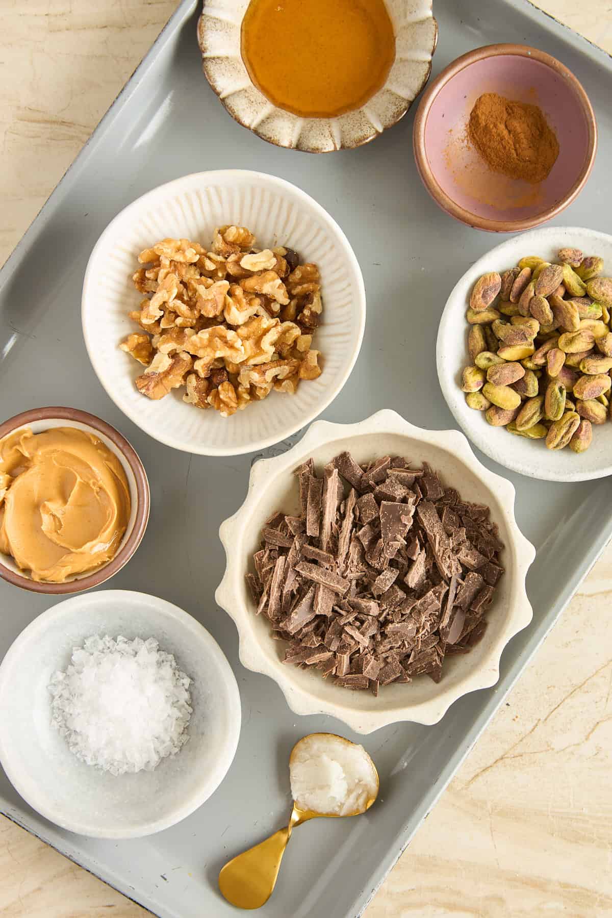 A gray tray holds bowls of chopped chocolate, walnuts, pistachios, peanut butter, sea salt, honey, cinnamon, and a spoon with coconut oil on a light-colored surface.
