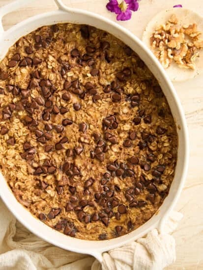 A baked oatmeal casserole topped with chocolate chips in a white oval dish, set on a light surface with a beige cloth, small bowl of walnuts, and purple flowers nearby.