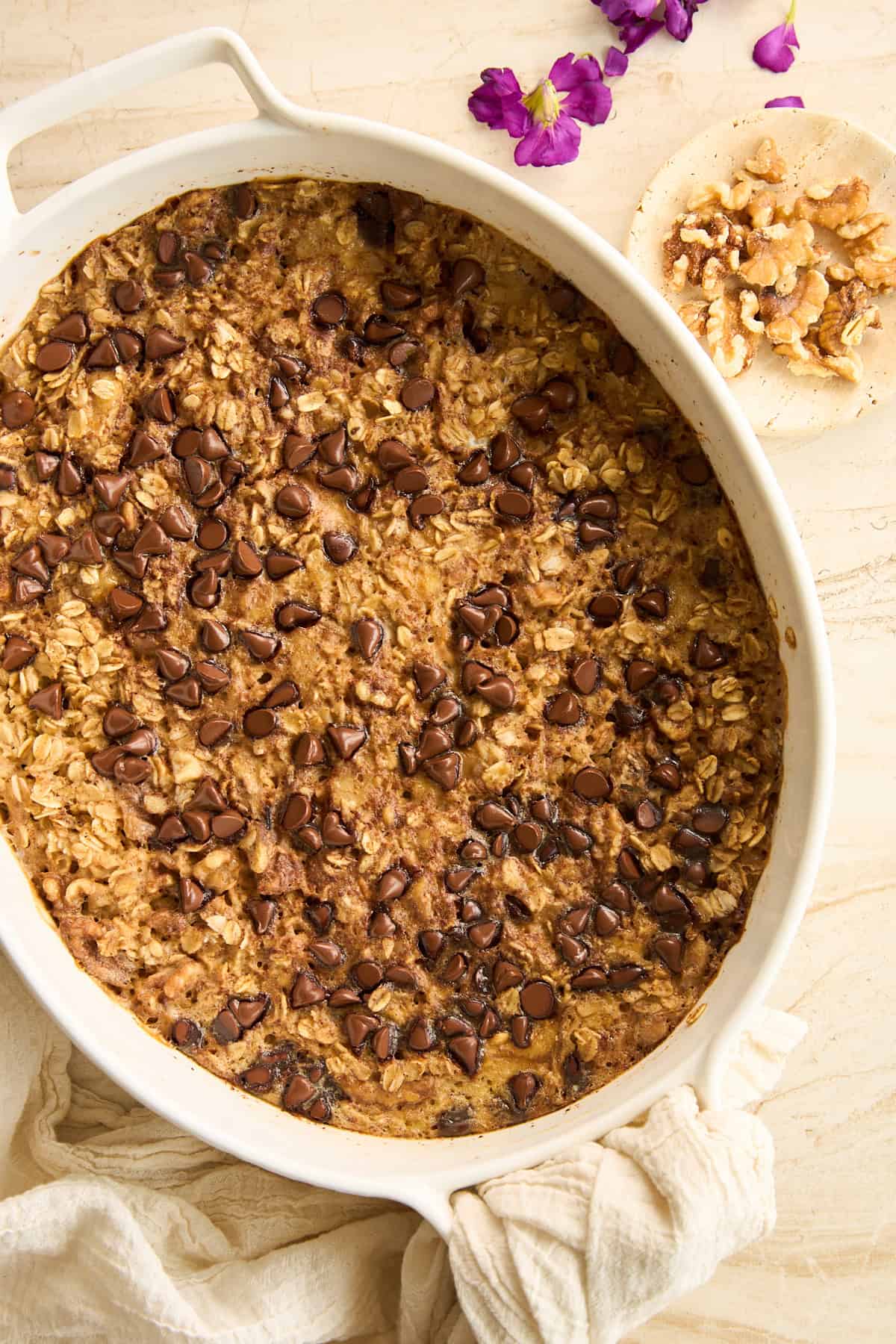 A baked oatmeal casserole topped with chocolate chips in a white oval dish, set on a light surface with a beige cloth, small bowl of walnuts, and purple flowers nearby.