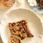 A white plate with a square slice of baked oatmeal topped with chocolate chips, a fork holding a small bite, a glass pitcher of honey, white flowers, and part of another baked oatmeal slice on a gray plate nearby.