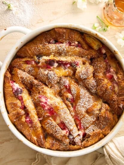 A round baking dish filled with golden-brown croissant bread pudding, dotted with red berries and dusted with powdered sugar. A beige cloth, white flowers, and a small jar of syrup surround the dish.