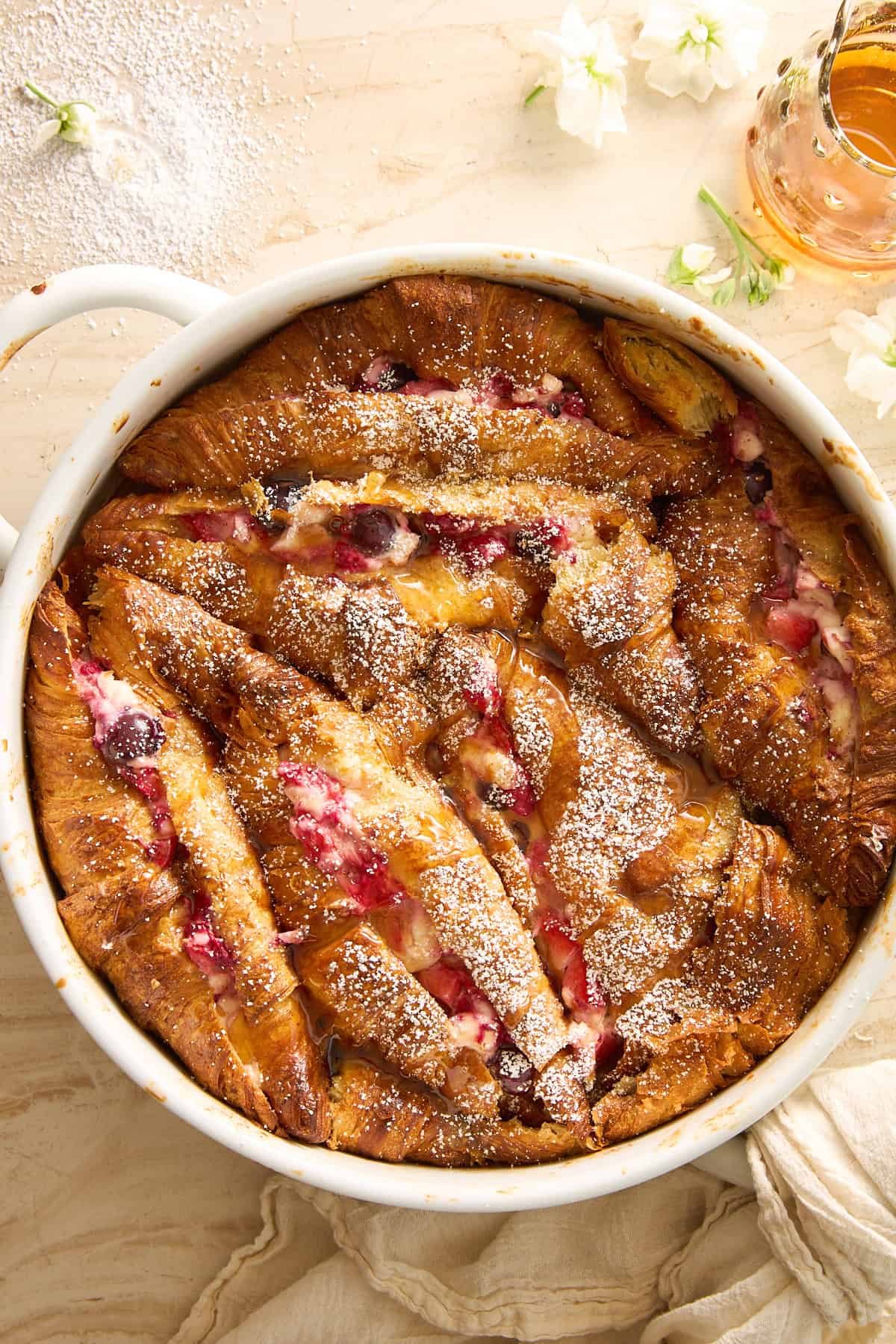 A round baking dish filled with golden-brown croissant bread pudding, dotted with red berries and dusted with powdered sugar. A beige cloth, white flowers, and a small jar of syrup surround the dish.