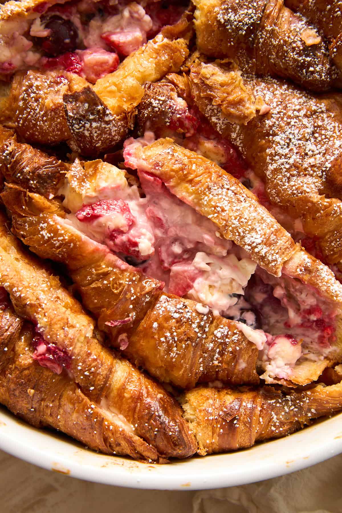 Close-up of a baked dessert made with croissants, mixed berries, and cream, dusted with powdered sugar and served in a round white dish. The croissants are golden and slightly crispy on top.