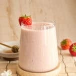 A glass of creamy strawberry smoothie sits on a round stone coaster, garnished with a whole strawberry. Nearby are two strawberries, a plate with a honey dipper, and white flowers, all on a tiled surface.