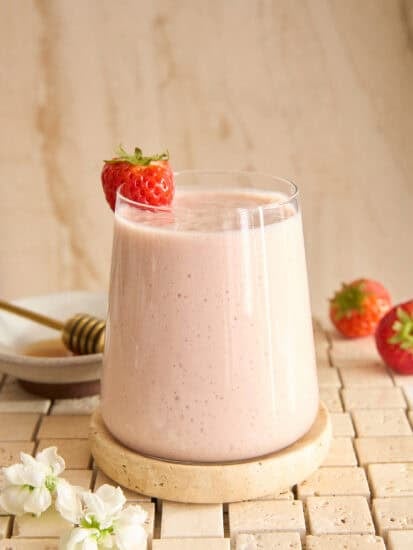A glass of creamy strawberry smoothie sits on a round stone coaster, garnished with a whole strawberry. Nearby are two strawberries, a plate with a honey dipper, and white flowers, all on a tiled surface.