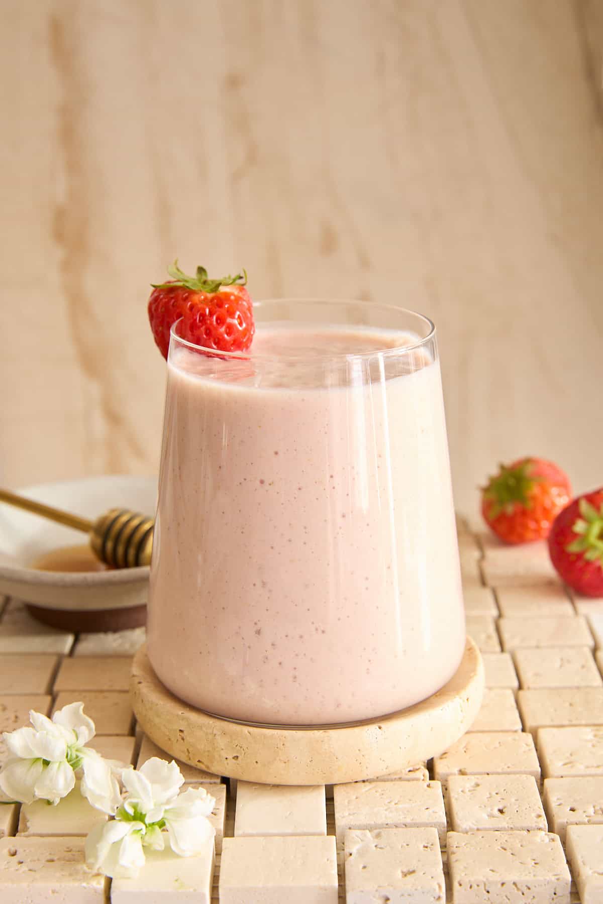 A glass of creamy strawberry cottage cheese smoothie sits on a round stone coaster, garnished with a whole strawberry. Nearby are two strawberries, a plate with a honey dipper, and white flowers, all on a tiled surface.