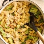 A pot of rotini pasta mixed with pieces of chicken and broccoli, topped with grated cheese, sits on a light countertop. A black serving spoon rests in the pot, and a bowl of red pepper flakes is nearby.