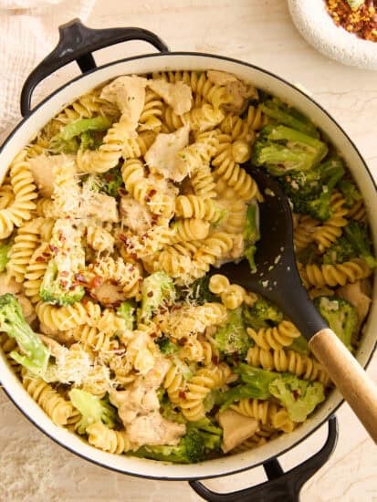 A pot of rotini pasta mixed with pieces of chicken and broccoli, topped with grated cheese, sits on a light countertop. A black serving spoon rests in the pot, and a bowl of red pepper flakes is nearby.