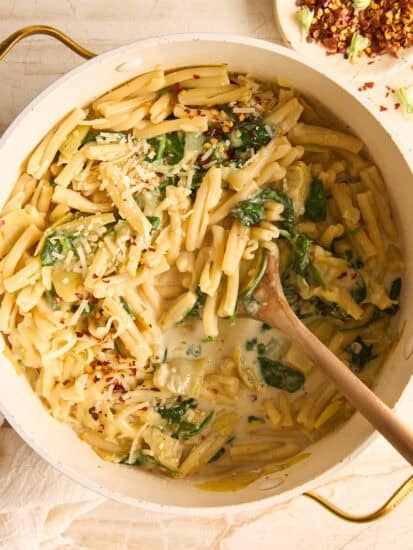 A pot of creamy pasta with spinach and artichokes, topped with grated cheese. A wooden spoon rests in the pasta, and a plate with red pepper flakes is visible in the background.