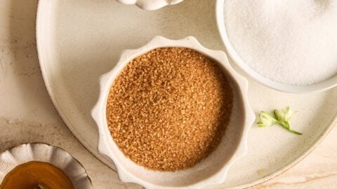 A top-down view of various sweeteners in small bowls on a beige surface, including honey with a dipper, brown sugar, white sugar, and a golden liquid, each with a spoon.
