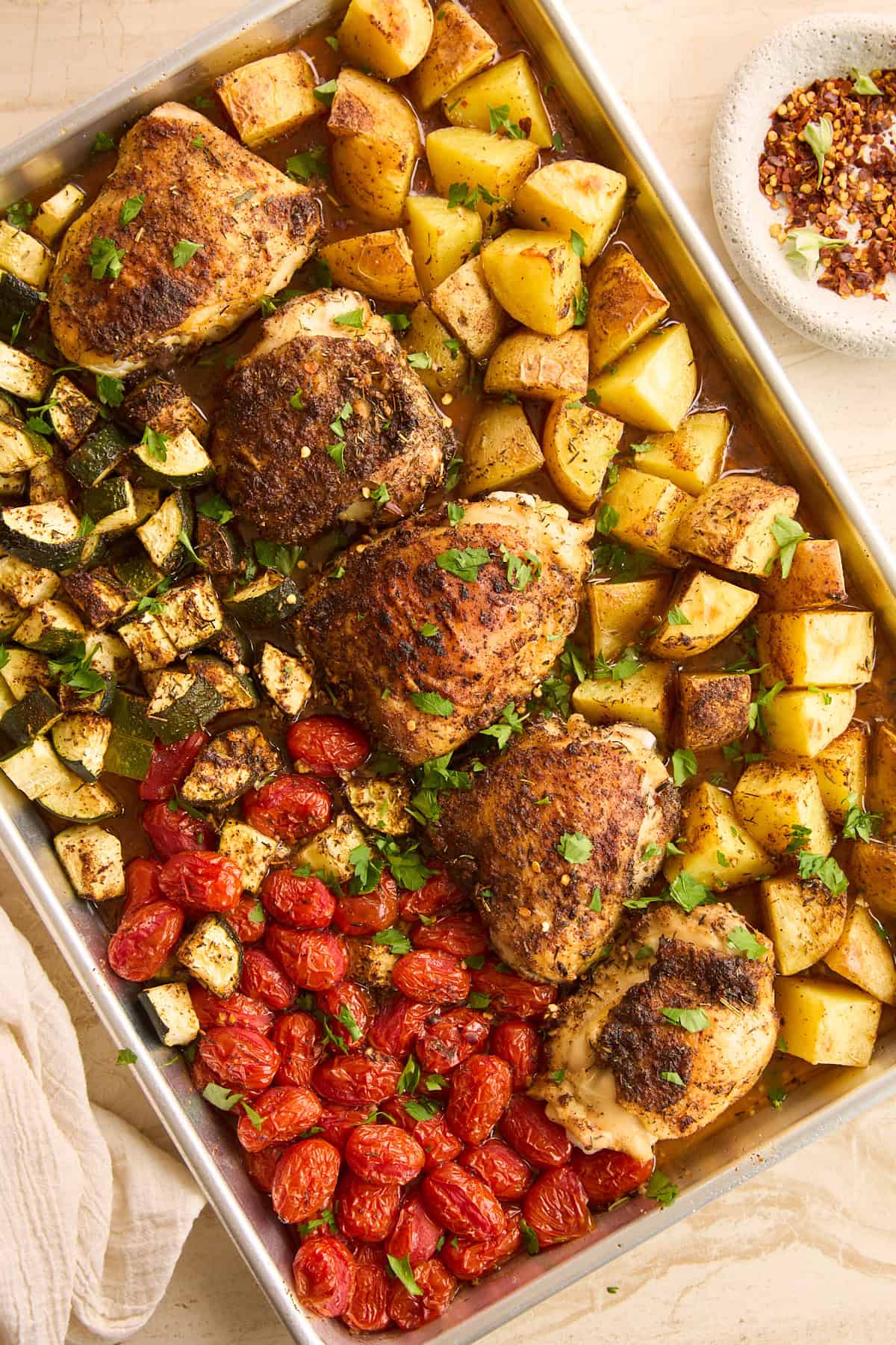 A sheet pan filled with roasted chicken thighs, golden potato chunks, diced zucchini, and cherry tomatoes, garnished with fresh herbs, next to a bowl of red pepper flakes.