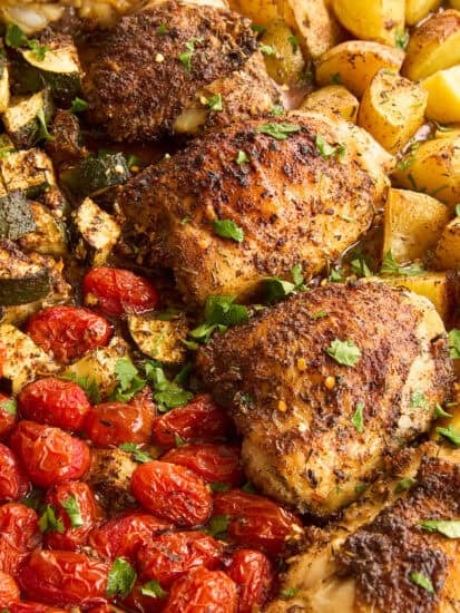 Seasoned roasted chicken thighs on a sheet pan with golden roasted potatoes, cherry tomatoes, and zucchini, garnished with chopped fresh herbs.